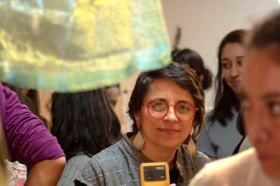 In a group of people Tania Perez-Bustos is centered in the frame, her attention is fixed on a piece of fabric that can be seen hanging in the foreground. 